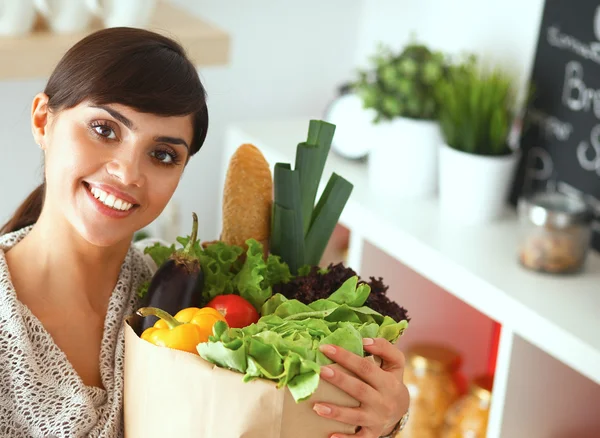 Mujer joven sosteniendo bolsa de la compra de comestibles con verduras de pie en la cocina —  Fotos de Stock