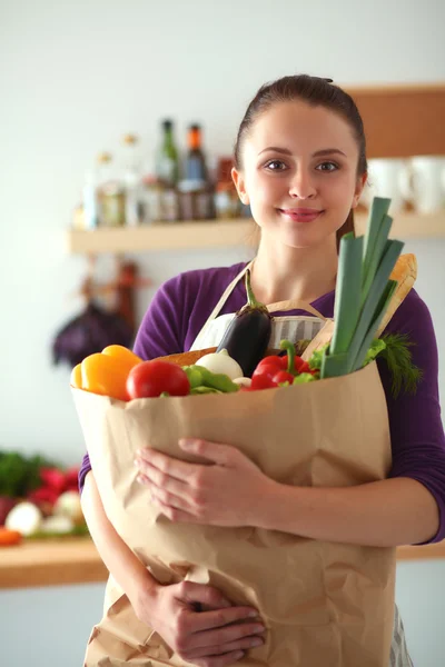 Mujer joven sosteniendo bolsa de la compra de comestibles con verduras de pie en la cocina —  Fotos de Stock