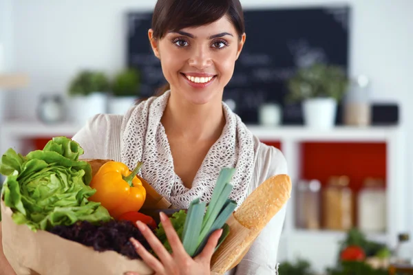 Mujer joven sosteniendo bolsa de la compra de comestibles con verduras de pie en la cocina —  Fotos de Stock