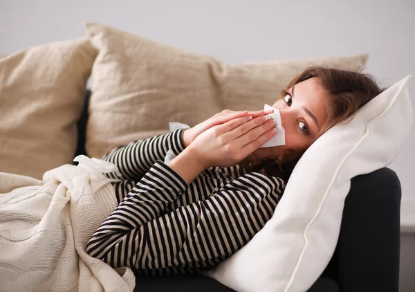 Retrato de una mujer enferma sonándose la nariz mientras está sentada en el sofá — Foto de Stock