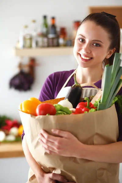 Mujer joven sosteniendo bolsa de la compra de comestibles con verduras de pie en la cocina —  Fotos de Stock