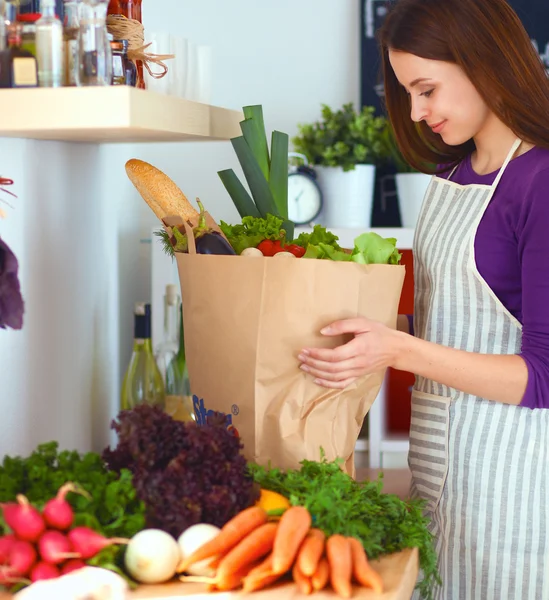 Mujer joven de pie en su cocina cerca del escritorio con bolsas de compras —  Fotos de Stock