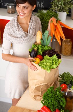 Sebzeli alışveriş poşeti taşıyan genç bir kadın mutfakta duruyor.