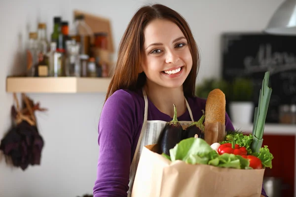 Mujer joven sosteniendo bolsa de la compra de comestibles con verduras de pie en la cocina — Foto de Stock