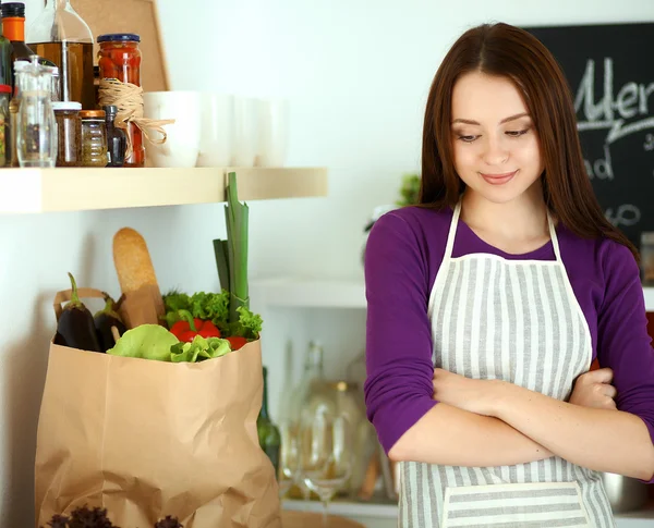 Mujer joven de pie en su cocina cerca del escritorio con bolsas de compras —  Fotos de Stock