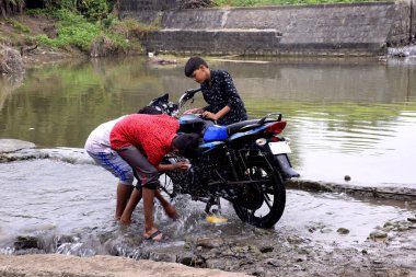 Kırsal çocuklar bisikletleri nehir suyuyla yıkıyor, doğal yeşillik arka planda.