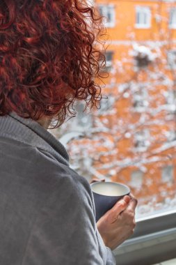Kızıl saçlı kadın pencereden bakarken bir fincan kahve içiyor.