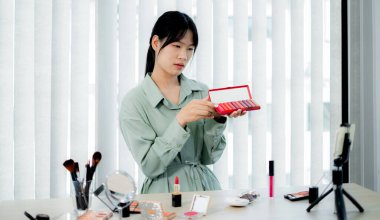 Güzel Asyalı kadın vlogger video kaydederken kozmetik makyaj ürünleri gösteriyor ve dijital akıllı telefonuyla güzellik blogu için tavsiye ediliyor.