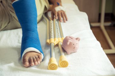 Adamın bacağı ameliyat sonrası yürümek için koltuk değneği kullanıyor. Kırık kemikler ve pembe domuz kumbarası..