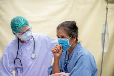 Yaşlı kadın, doktor sahra hastanesindeki hastanın öksürük belirtilerini inceliyor, ikisi de salgın hastalık nedeniyle maske takıyor..