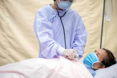 Yaşlı hastalar yatakta yatıyor, doktorlar akciğer fonksiyonlarını kontrol etmek için steteskop kullanıyor. İkisi de COVID salgını yüzünden maske takıyordu..