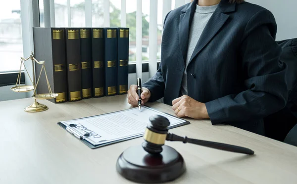 Kadın avukat elinde adalet terazisi ve tokmak çekiciyle büroda oturuyor. Müşterilere tavsiye vermek için iş anlaşmazlıklarında hukuk okuyun.
