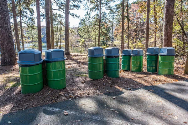 Eight green metal drum garbage cans with plastic lids in a row secured to posts at a picnic area alongside a parking lot in a park on a bright sunny day in autumn