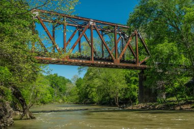 A fast moving river flowing underneath the old rusty iron train trestle with woodlands surrounding on a vibrant blue sky day in springtime clipart