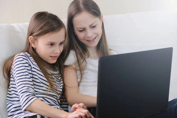 Twee schoolmeisjes zitten thuis op de bank emotioneel kijkend naar een laptop. — Stockfoto