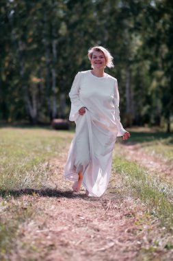 Beyaz elbiseli mutlu genç kadın yaz orman yolunda yürüyor.