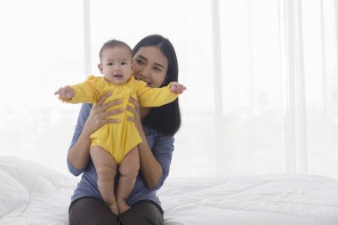 Asyalı bir anne mutlu bir şekilde bebeğiyle yatakta oynuyor..