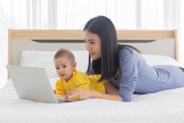 Asyalı bir anne yatakta dizüstü bilgisayarla ve bebeğiyle evde iş kavramının yanında çalışıyor..