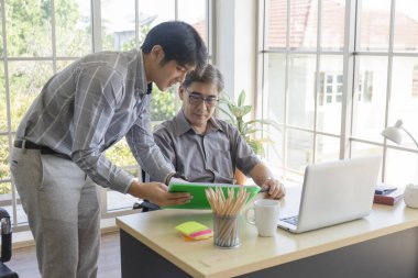 Orta yaşlı Asyalı bir adam oğluna bir ofiste masanın yanında duran bir iş öğretiyor..