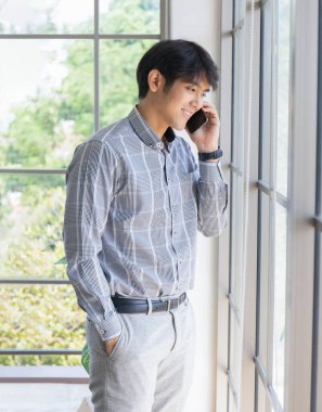 Genç bir Asyalı işadamı bir pencerenin önünde telefonla konuşuyor..