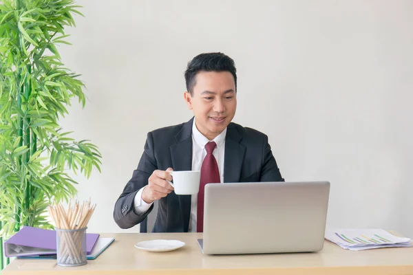 The manager of an Asian man sipping coffee on his desk in a radiant manner.