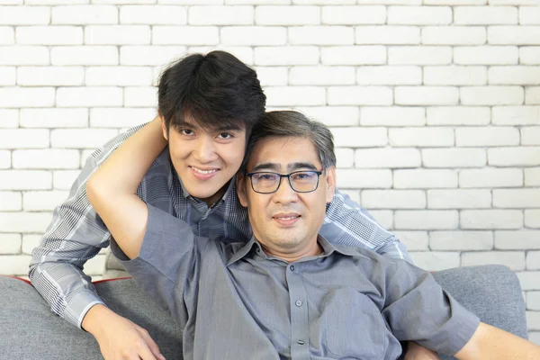 A middle-aged father and an Asian son hugged each other on the sofa with a happy smile.