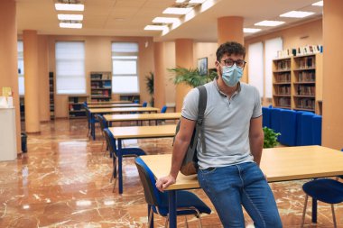 Maskeli çocuk arka planda boş kütüphaneyle kameraya bakıyor.