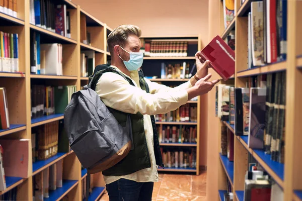 a young man with a mask is picking up a book in the university library