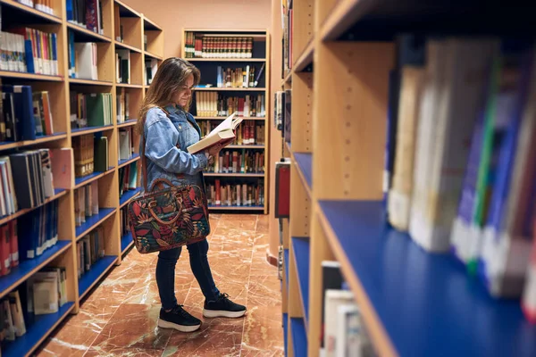 Genç kız bir kütüphanenin kitap rafları arasında bir kitaba bakıyor.