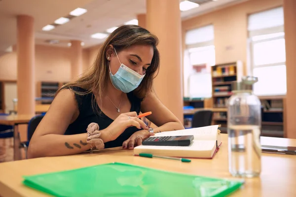 Üniversite kütüphanesinde cerrahi maskeli genç bir kadın okuyor.