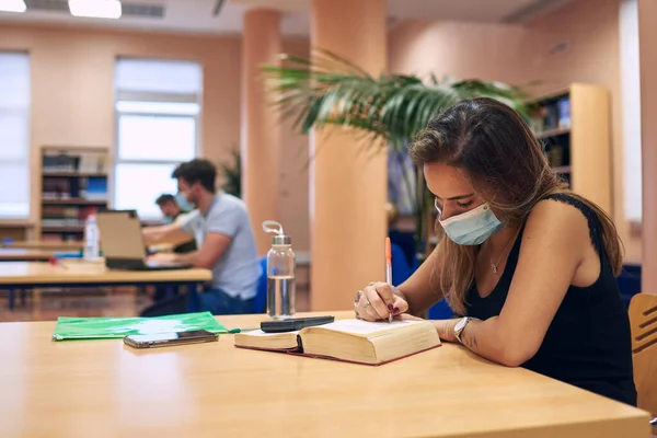 a girl in a mask studying in the library keeping a safe distance