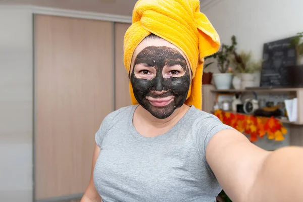 A woman with a cosmetic mask on her face made of clay, a towel on her head makes a selfie.
