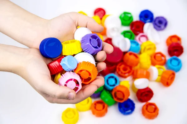 In children\'s palms there are plastic bottle caps against the background of other multi-colored plastic bottle caps. Ecology and recycling concept.