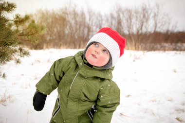 Kışın yeşil tulumlu ve parkta kırmızı şapkalı bir çocuk. Kışın kırmızı yanaklı bir çocuk.