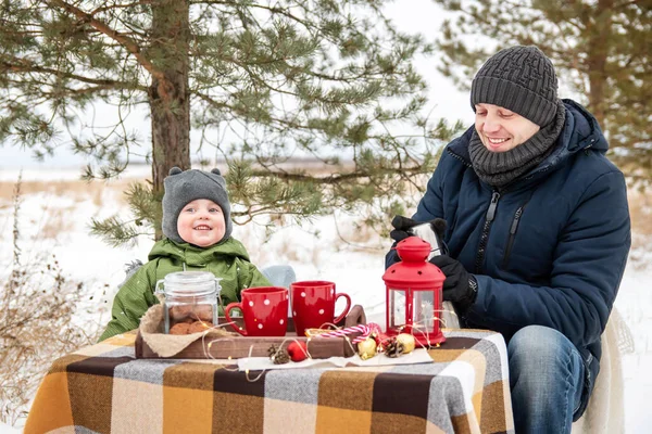 森林里的冬季野餐。爸爸和儿子坐在桌旁，喝热水瓶里的热茶. — 图库照片