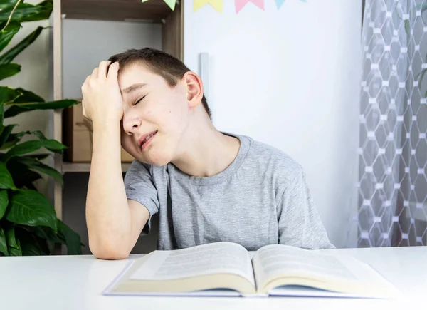 The boy doesn\'t want to read the book. A boy in a gray T-shirt sits at a white table at the open book and holds his hand behind his head