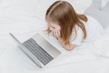 Uzun saçlı küçük bir kız yatağa uzanıyor ve dizüstü bilgisayarına bakıyor. Kavramsal çocuk ve aletler, internet, bağımlılık