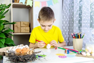 Sarı tişörtlü bir çocuk bir masaya oturur ve renkli kalemlerle beyaz bir yumurta boyar. Paskalya hazırlığı kavramı