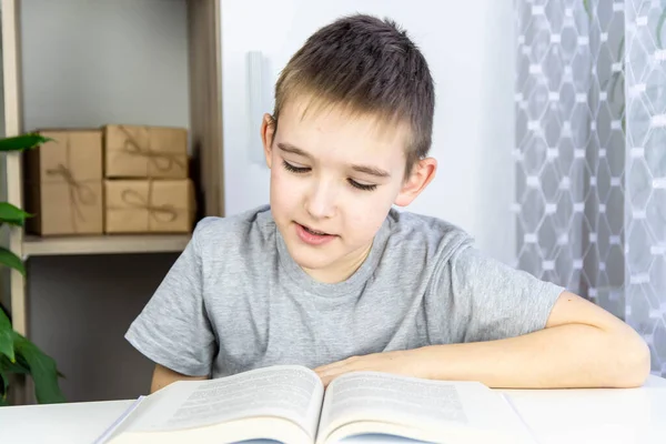 A boy reads a book. A schoolboy in a gray T-shirt at home at a white table looks into a book