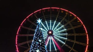 Dönme dolap dönüyor ve yeni yıl ağacı Kharkiv şehir merkezindeki parka yakın görünüyor. Gece renkli parlak aydınlatma. Mevsimlik tatil dekorasyonu