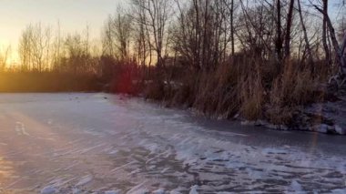Koyu ağaçlar siluetleri, uzun gölgeler ve açık gökyüzü ile donuk yansıtıcı gölde turuncu gün batımı soldan sola doğru hareket ediyor.