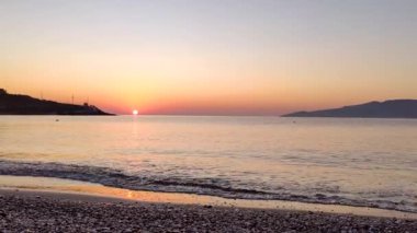 Yunanistan 'ın Atina kenti yakınlarındaki çakıl taşlı sahil kıyısında açık gökyüzü ufkunda parlayan turuncu gün doğumu güneşi. Yaz seyahati Sabahları sakin deniz kıyıları