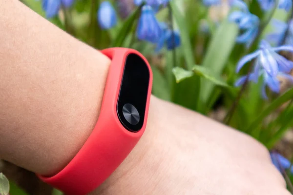 Fitness tracker with red band on wrist close-up in spring blue flowers. Outdoor activity wellness monitor gadget on female arm with focus blurred blooming background