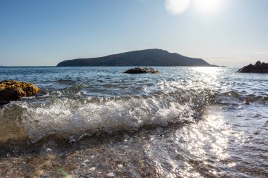 Güneş beyaz dalgaların üzerinde parlıyor ve kıyıya vuruyor. Akdeniz 'de kayalar, berrak su ve güneş ışığı. Atina yakınlarında Yunanistan 'ı gezmek. Yaz deniz kenarı doğası. Mavi filtre