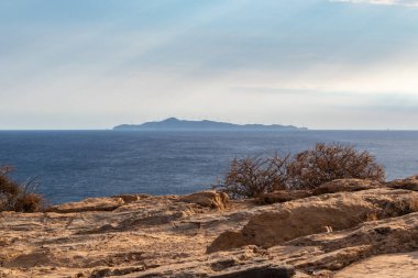 Yunanistan 'ın kayalık kıyılarında bulutların üzerinde parlayan güneş ışınları. Akdeniz 'de deniz manzarası ve gün batımında ada manzarası. Perspektif görünümü