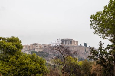 Akropol tepesi (Parthenon, Propylaea, Tapınaklar, Atticus 'un Odeonu) yeşil ağaç dalları arasından görülür. Bulutlu bir günde Atina Filopappou Tepesi 'nden kent merkezindeki tarihi eser