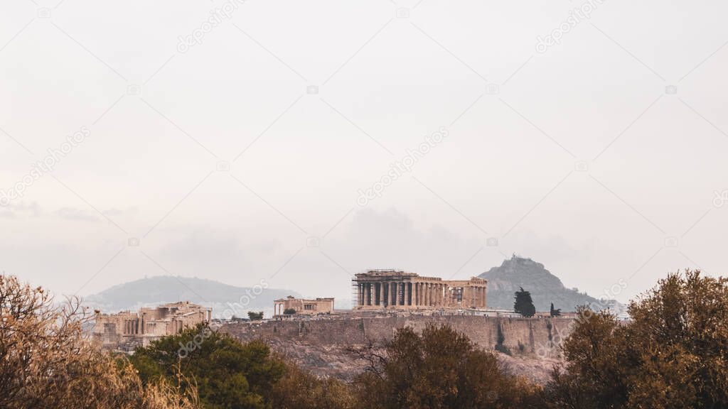 Acropolis (Parthenon, Propylaea, Temples) and Mount Lycabettus in summer bushes. Athens ancient historical landmark from Filopappou Hill on cloudy sky. Warm colors