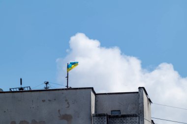 Ukrayna ulusal bayrağı, yüksek Sovyet mimarisinin çatısında rüzgarda sallanıyor. Mavi gökyüzü ve manzaralı beyaz bulutlar arka planda çok katlı bina.