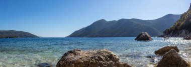 Güneşli çakıl taşlı plajda, kristal berrak gök mavisi suyla büyük kayanın manzarası. Yunanistan 'daki Lefkada adasının kıyısında. İyon Denizi 'ne yaz canlı doğa yolculuğu