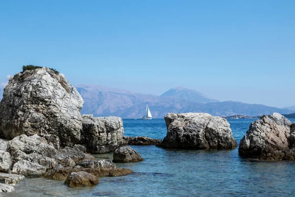 Kıyıda mavi berrak suları, beyaz yelkenli yatı ve ufukta dağları olan büyük kayalar, Yunanistan 'daki Lefkada adası. İyon Denizi 'ne yaz canlı doğa yolculuğu